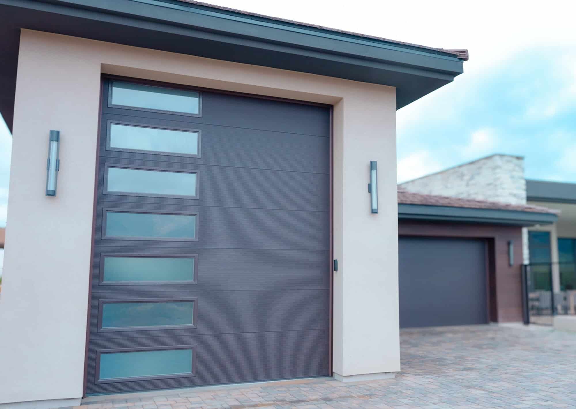 A contemporary, charcoal-gray garage door with a textured, horizontal-panel design. It features a vertical column of seven rectangular, frosted glass windows on the left side. The door is framed by off-white stucco walls and illuminated by two sleek, vertical light fixtures on either side. The driveway is paved with multi-toned interlocking stones, and a similar dark garage door is visible in the background against a modern house with stone and wood accents.