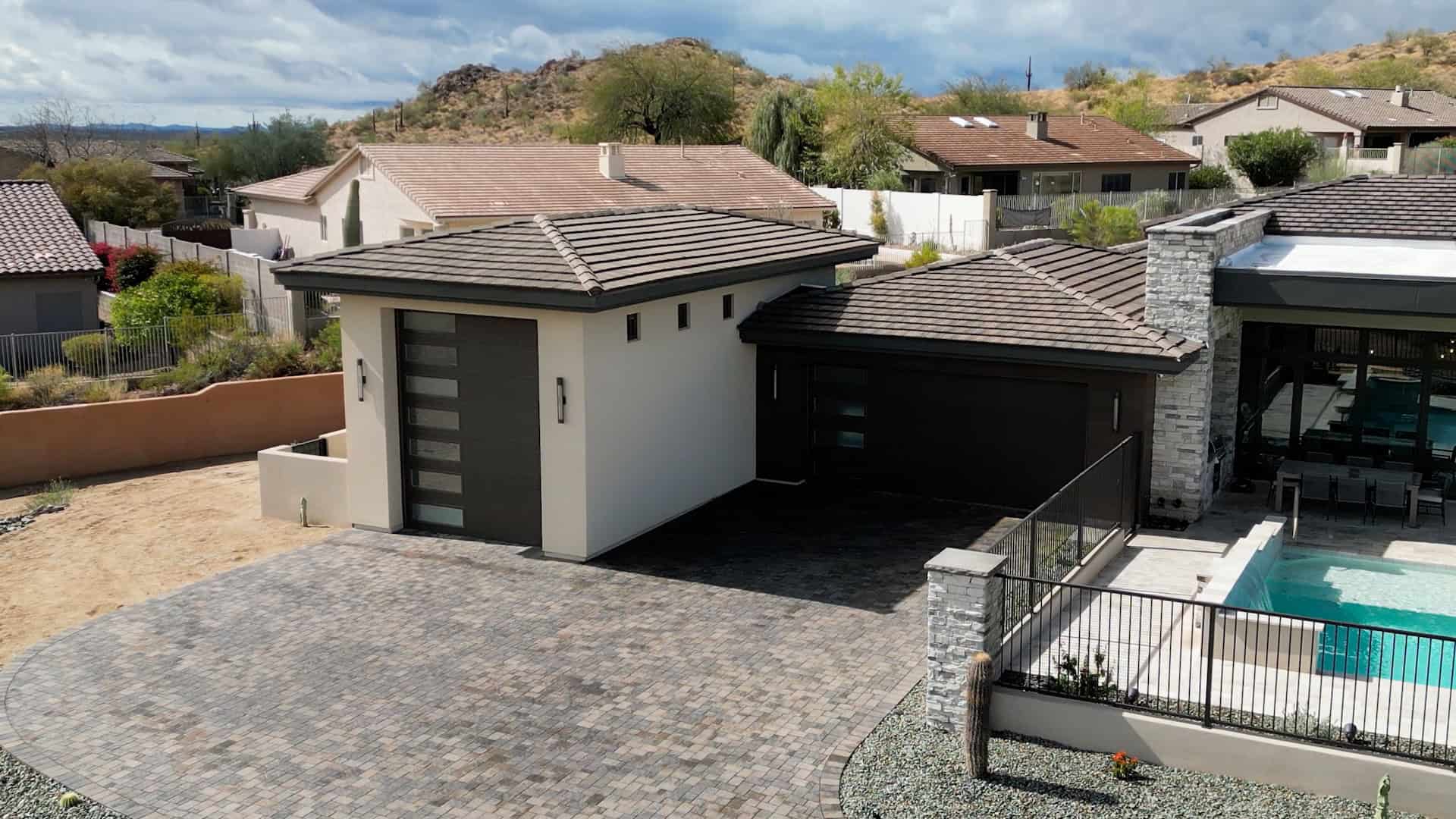 An aerial, high-angle view shows a modern desert home featuring a light-tan stucco exterior and a dark, flat-tiled roof. The property includes a detached single-car garage and an attached two-car garage, both equipped with dark charcoal doors that have vertical stacks of frosted windows. A large driveway made of multi-toned gray and tan stone pavers connects the structures. To the right, a contemporary outdoor living area is visible, featuring a patio with a dining set, a swimming pool with a stone water feature, and a black metal fence. The background reveals a hilly, arid landscape with sparse vegetation and neighboring residential homes under a cloudy sky.
