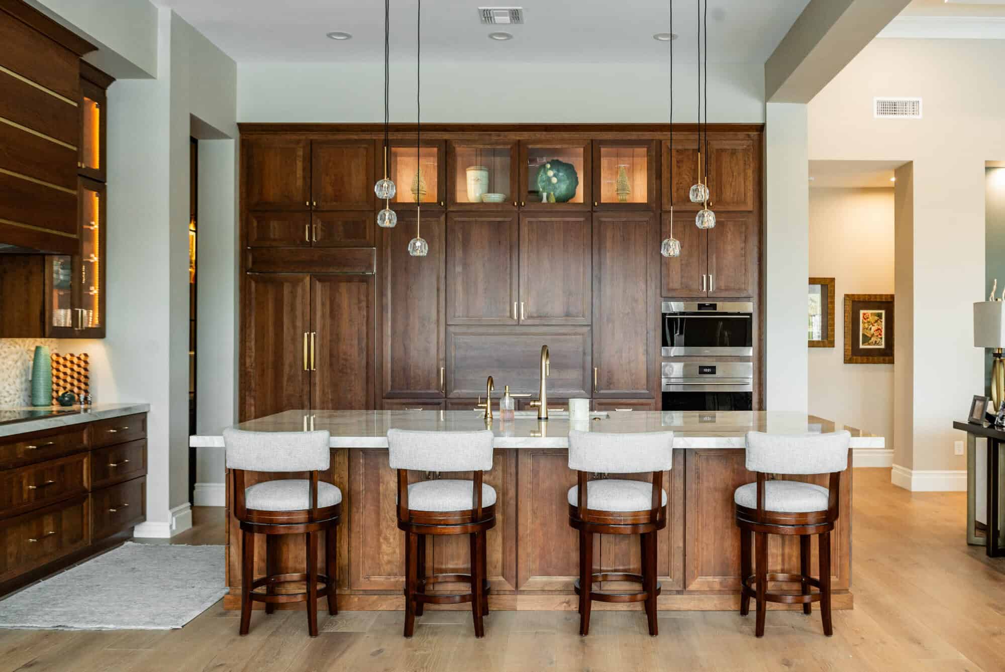 A luxurious, modern kitchen features rich, dark wood cabinetry and a large central island with a white marble countertop. Four light-gray upholstered barstools with dark wood legs are lined up along the island. Above the island hang three elegant gold pendant lights, each featuring a cluster of four glowing glass globes. The background reveals a matching dark wood pantry and upper cabinets with glass-fronted top sections that are internally lit, displaying glassware. A white marble backsplash complements the countertops, and a gold faucet serves as a central accent on the island. To the right, a stainless steel double wall oven is integrated into the cabinetry, and a glimpse of an adjacent dining area with a wooden console table and framed artwork is visible.