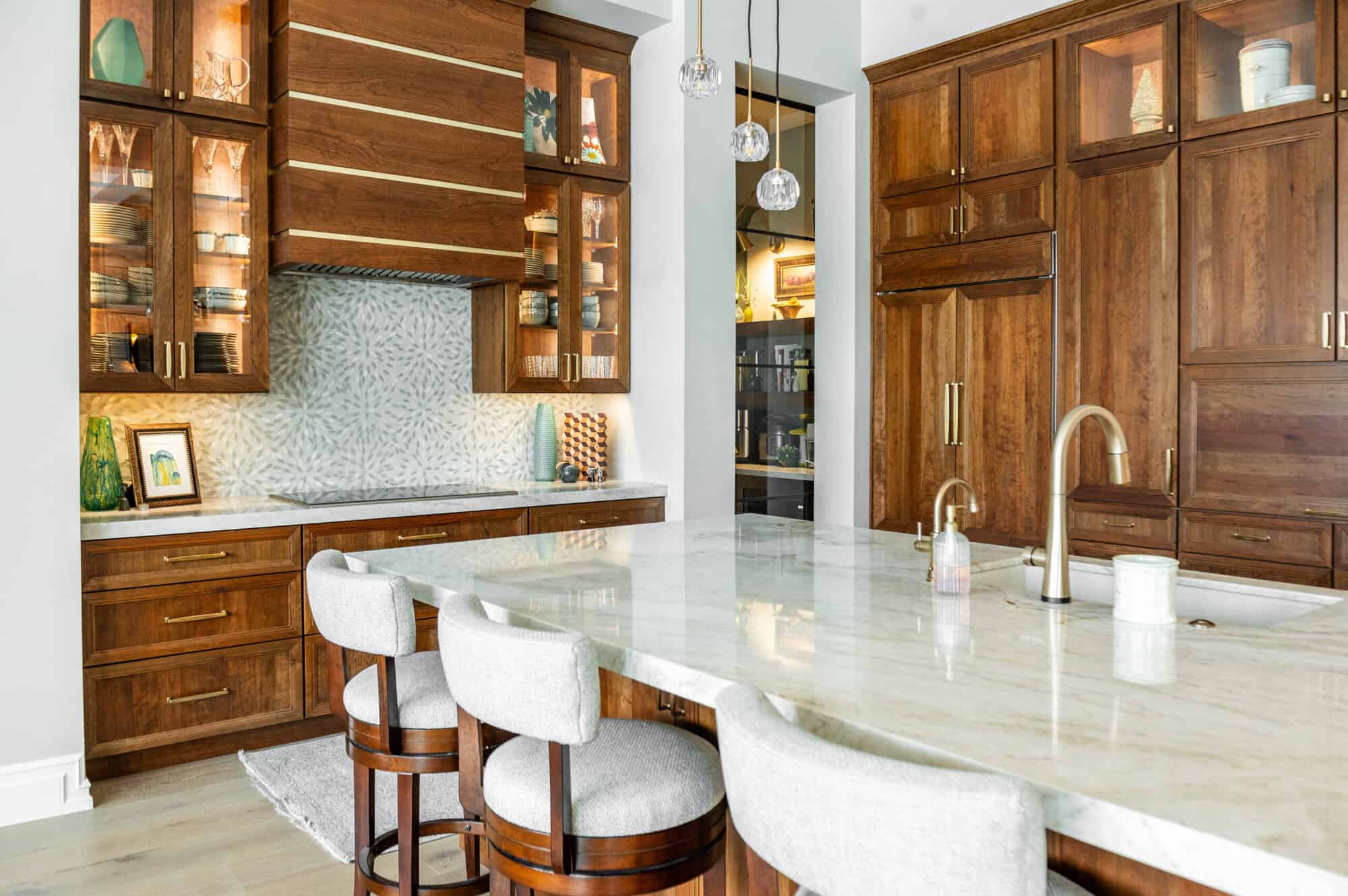 A luxurious modern kitchen centered around a large white marble island with a gold faucet and four light-gray upholstered barstools. The back wall features floor-to-ceiling warm-toned wood cabinetry, including glass-front upper cabinets with internal lighting that displays decorative items. To the left, a secondary workspace includes a patterned tile backsplash and a large wood-paneled range hood with gold horizontal accents. Three delicate glass globe pendant lights hang over the island, and the room is finished with light-colored wide-plank wood flooring.