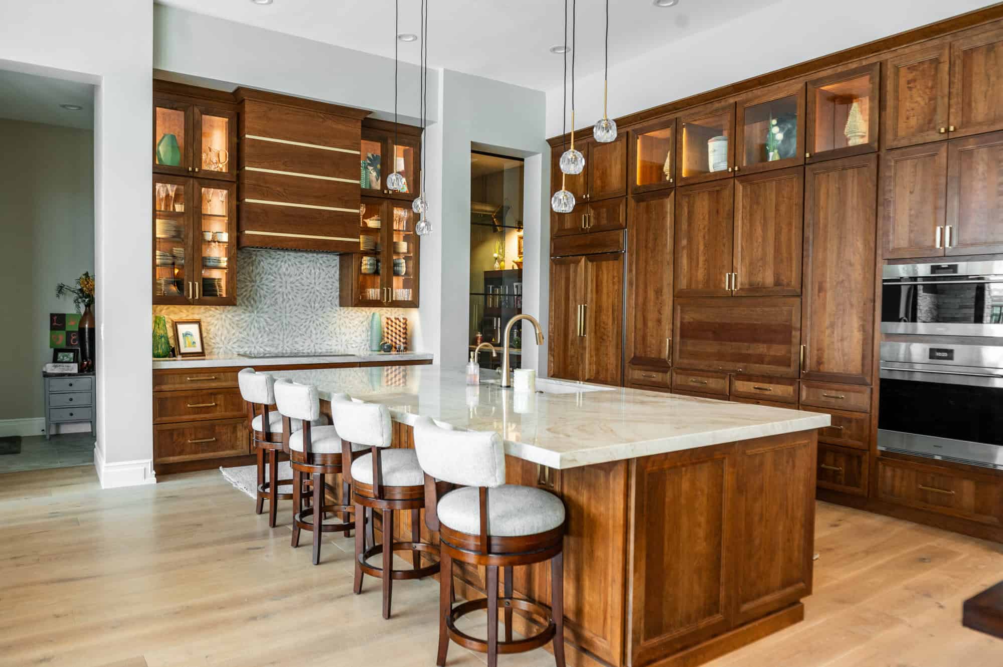 A luxurious modern kitchen centered around a large white marble island with a gold faucet and four light-gray upholstered barstools. The back wall features floor-to-ceiling warm-toned wood cabinetry, including glass-front upper cabinets with internal lighting that displays decorative items. To the left, a secondary workspace includes a patterned tile backsplash and a large wood-paneled range hood with gold horizontal accents. Three delicate glass globe pendant lights hang over the island, and the room is finished with light-colored wide-plank wood flooring.