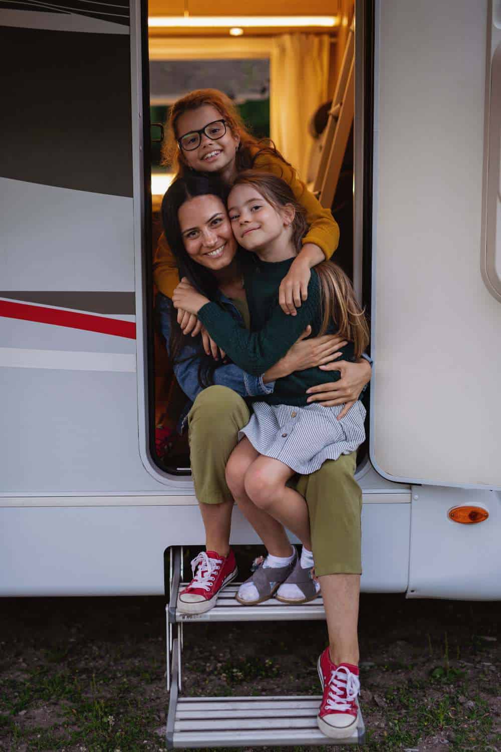 A woman and two young girls are smiling and hugging while sitting in the open doorway of a white recreational vehicle (RV). The woman sits on the threshold wearing olive green pants and red sneakers, while one girl in a dark sweater and striped skirt sits on her lap. The second girl, wearing glasses and a yellow sweater, leans over them from inside the van. A set of metal pull-down steps leads from the doorway to the ground.