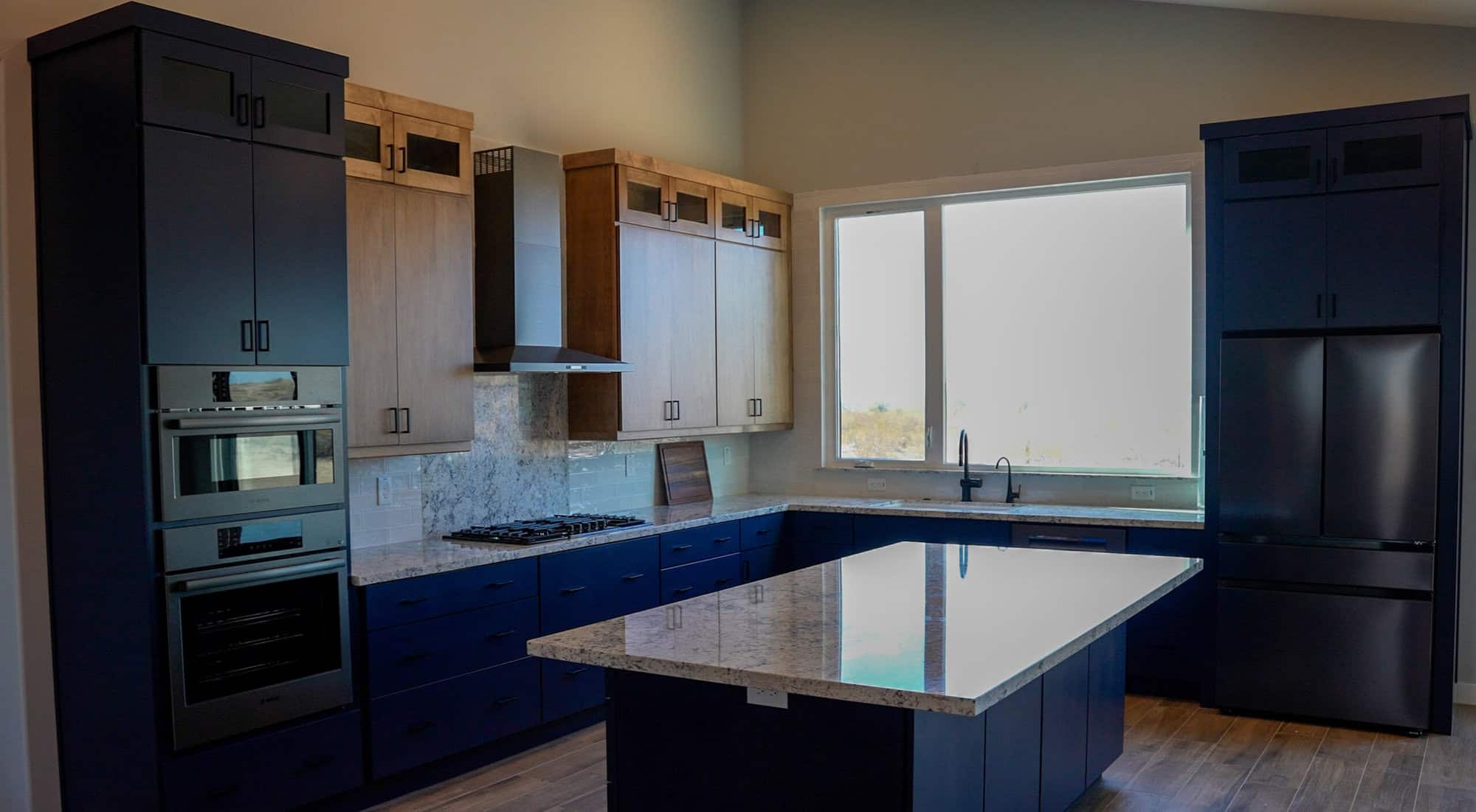 The kitchen features navy blue lower cabinetry and tall appliance towers paired with natural wood upper cabinets that include frosted glass inserts at the top. The countertops and large central island are a light, speckled granite or quartz, complemented by a grey marble-patterned backsplash behind the gas cooktop. High-end stainless steel appliances are integrated throughout, including a double wall oven and a large French-door refrigerator. A wide window above the sink looks out onto a bright landscape, filling the room with natural light.