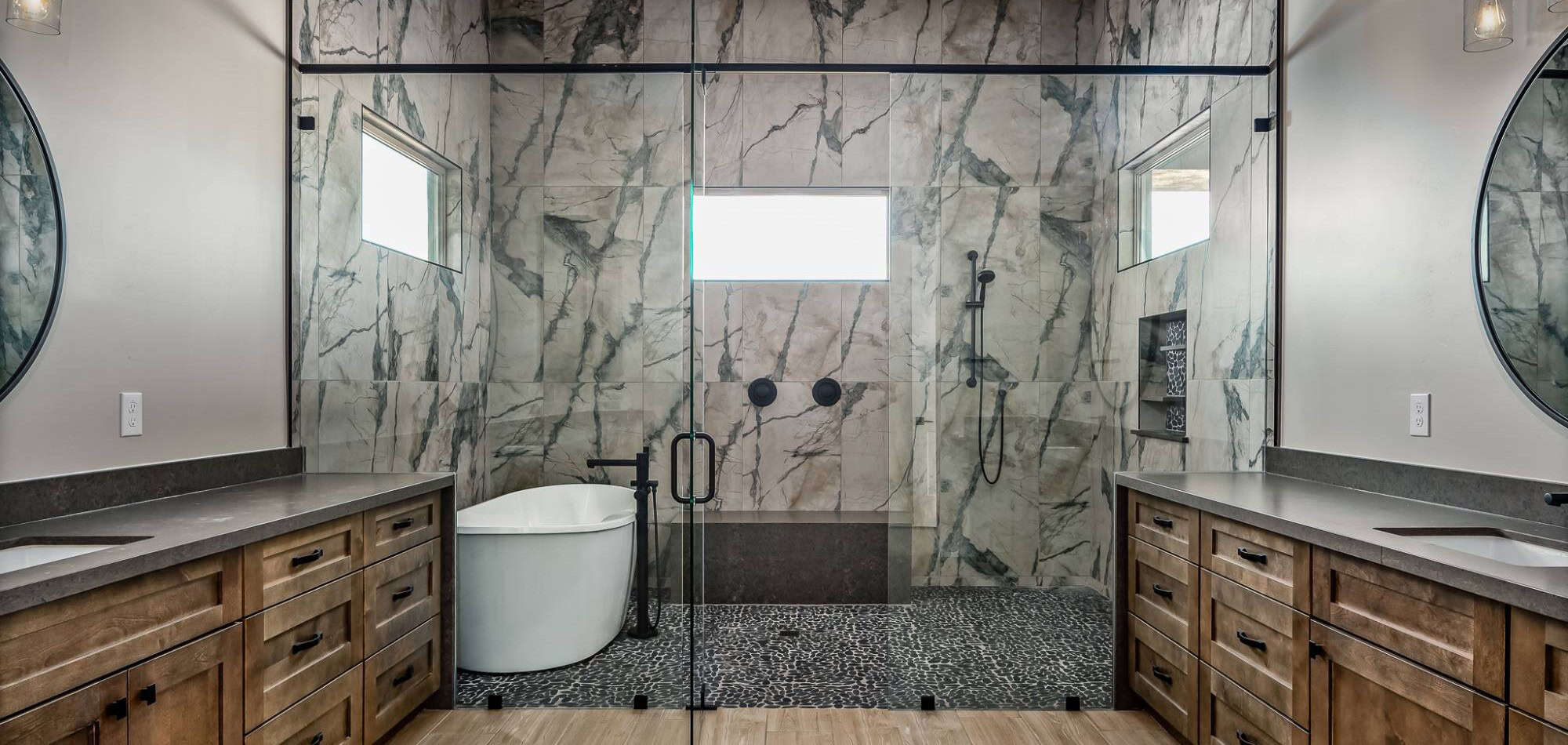 An expansive luxury primary bathroom features a large, glass-enclosed "wet room" area containing both a freestanding white soaking tub and a walk-in shower. The wet room is finished with dramatic, large-format marble tiles with heavy grey veining on the walls and a dark grey pebble stone floor. The room is symmetrical, with two matching wood vanities topped with dark grey countertops and black hardware on either side of the glass enclosure. Inside the wet room, black fixtures include a floor-mounted tub filler and a wall-mounted shower head with a handheld wand. Small horizontal windows are placed high on the walls, allowing for natural light while maintaining privacy.