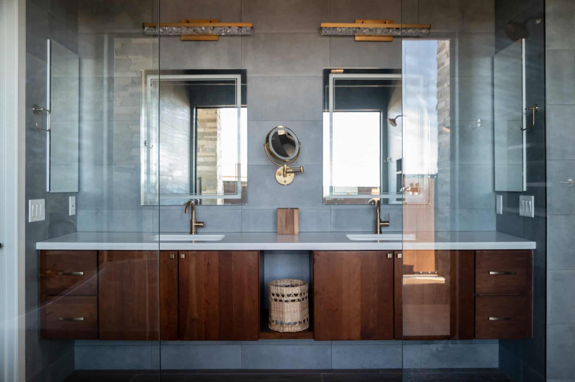 A wide shot of a modern, symmetrical bathroom vanity. Two rectangular mirrors with backlit edges are mounted against a dark gray tiled wall. Above each mirror is a horizontal, textured gold light fixture. A smaller, circular brass makeup mirror is mounted between the two larger mirrors. The vanity features a white countertop with two undermount sinks and brass faucets. Below the counter, the cabinetry is made of warm, dark wood with vertical grain. A central open shelf under the counter holds a woven basket. The entire scene is viewed through a glass shower partition, reflecting the room behind the camera.