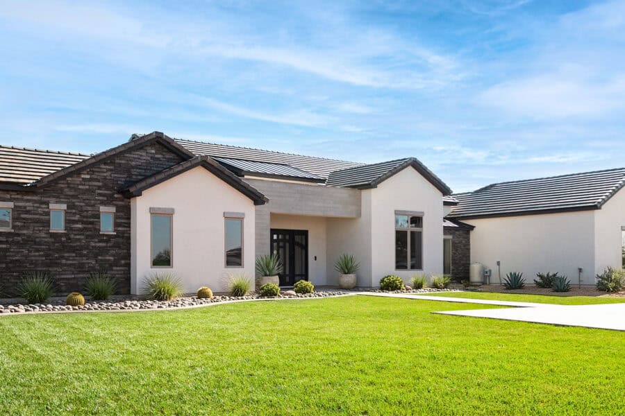 A wide, exterior shot of a large, modern single-story home under a bright blue sky with wispy clouds. The house features a mix of architectural textures, including dark gray stacked stone on the left wing and smooth off-white stucco on the central and right sections. The roof is composed of dark gray tiles with multiple gabled peaks. A vibrant, neatly manicured green lawn occupies the foreground, bordered by a desert-style landscape bed filled with smooth gray river rocks, small ornamental grasses, and barrel cacti. A concrete walkway leads toward a recessed front entryway with a dark double door. Large, minimalist windows with gray frames are scattered across the facade, and the overall aesthetic is clean and contemporary.