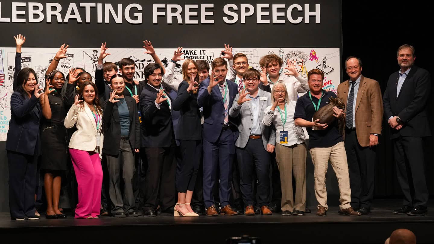 A diverse group of people poses on a stage under a sign that reads "CELEBRATING FREE SPEECH." Many are smiling and making an L-shaped hand gesture, with illustrated posters in the background.