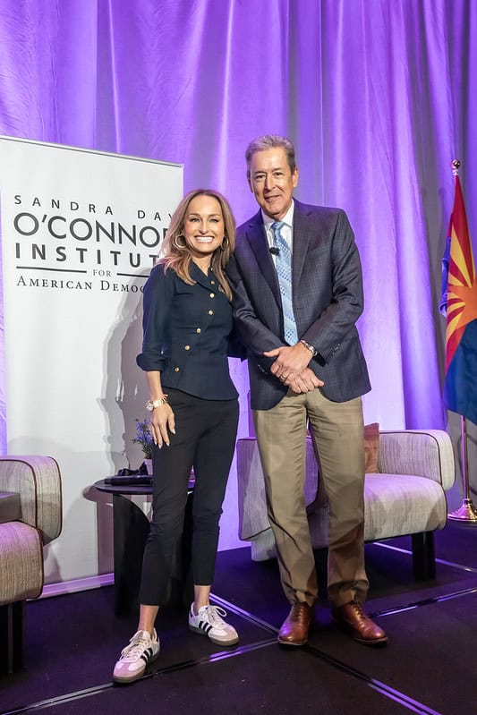 Two people stand and smile on a stage in front of a "Sandra Day O’Connor Institute for American Democracy" banner, with chairs and an Arizona state flag in the background. The lighting is bright with purple curtains.
