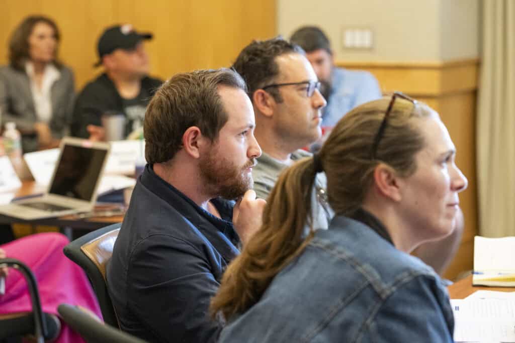Several adults sit attentively in a classroom or seminar setting, focusing on something out of view. Some have notebooks and laptops on the tables in front of them, actively engaging in Civics Education.