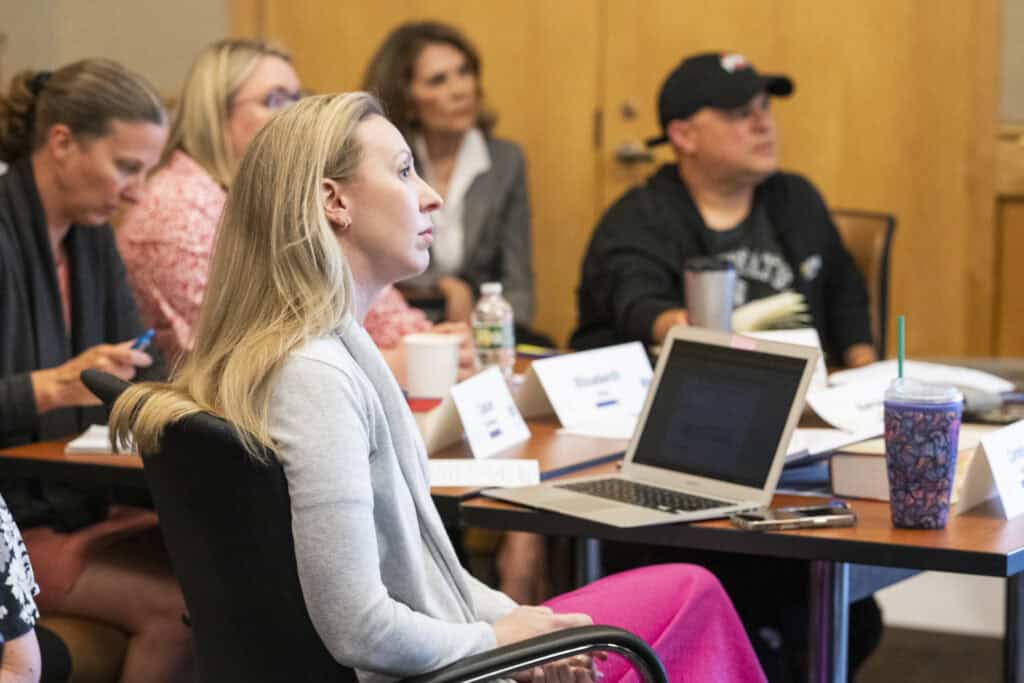 A group of people sit attentively around a conference table, engaging in a Civics Education discussion. A woman in the foreground looks forward with focus, with an open laptop and papers in front of her. Name cards are on the table.