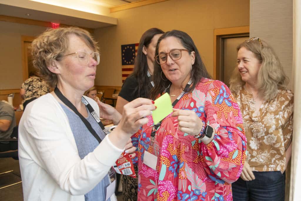 Four women stand together in a meeting room, smiling and holding colorful sticky notes during a Civics Education workshop. One woman wears glasses and a white cardigan, another wears a bright patterned top, while others look on cheerfully in the background.