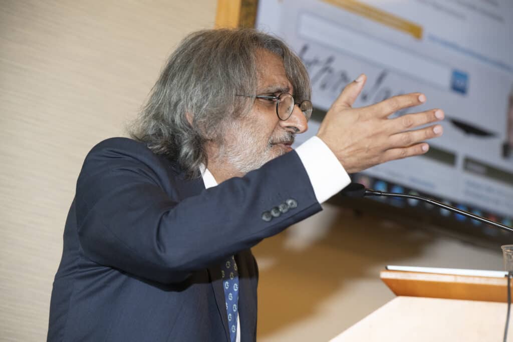 An older man with gray hair and glasses, wearing a suit and tie, gestures with his hand while speaking about Civics Education at a podium. A screen with text and images is visible in the background.