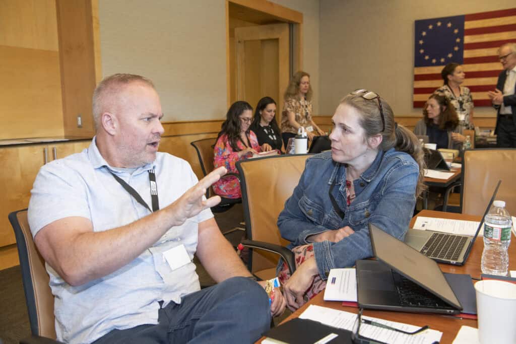 Two people sit at a conference table, engaged in conversation about Civics Education, with laptops, notebooks, and water bottles in front of them. Other attendees are in the background, and a U.S. flag hangs on the wall.