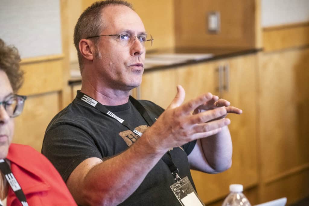 A man wearing glasses and a black t-shirt gestures while speaking about civics education in a discussion, seated indoors with a conference badge around his neck. Another person in a red jacket is partially visible beside him.