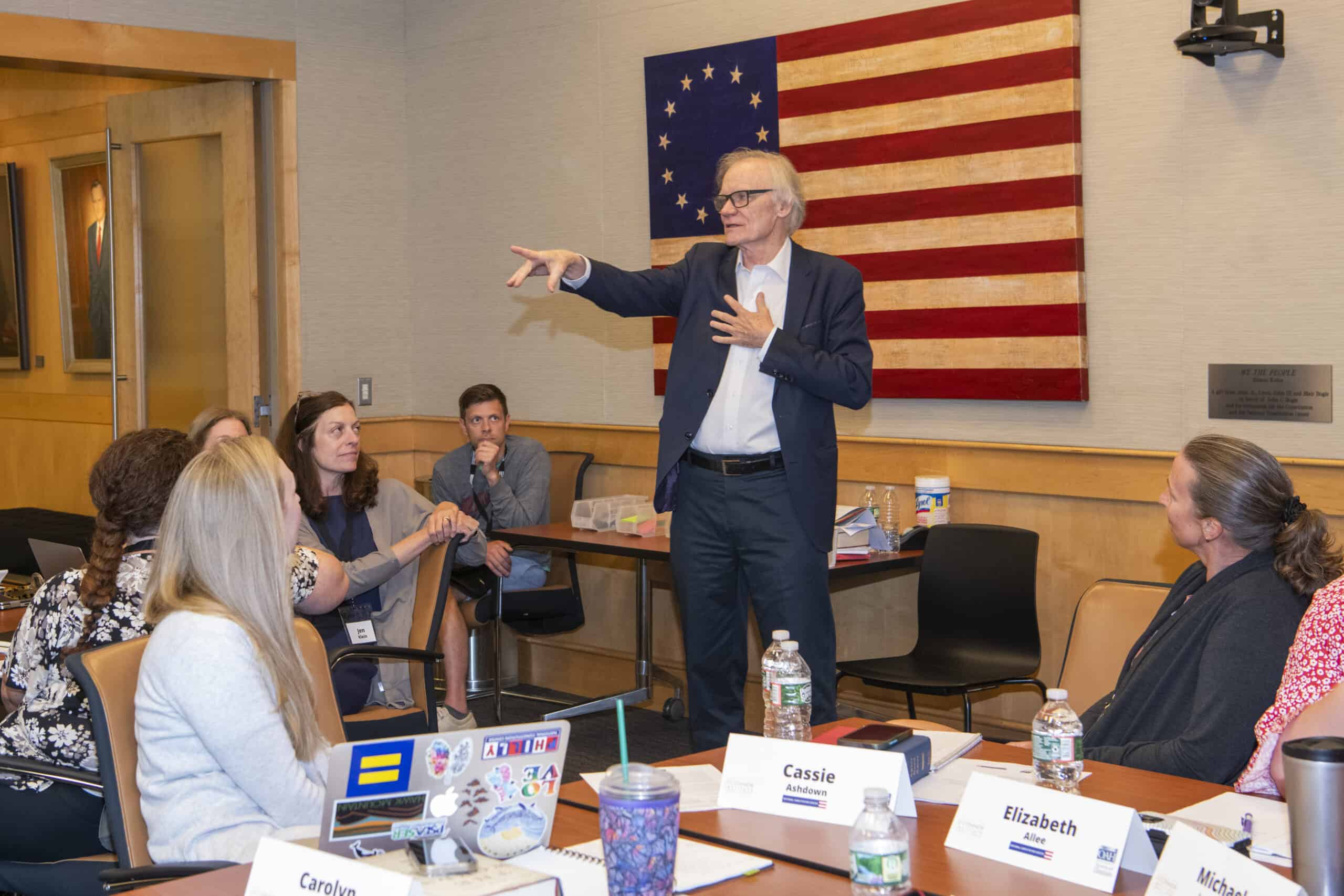 A man in a suit stands and gestures while speaking to a seated group in a meeting room, leading a civics education discussion with an American flag on the wall and name tags visible on the tables.