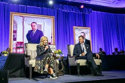 A woman and a man sit on stage in armchairs, speaking into microphones. Behind them are two large framed portraits and a blue curtain with stage lights. A table with people and flowers is visible in the background.