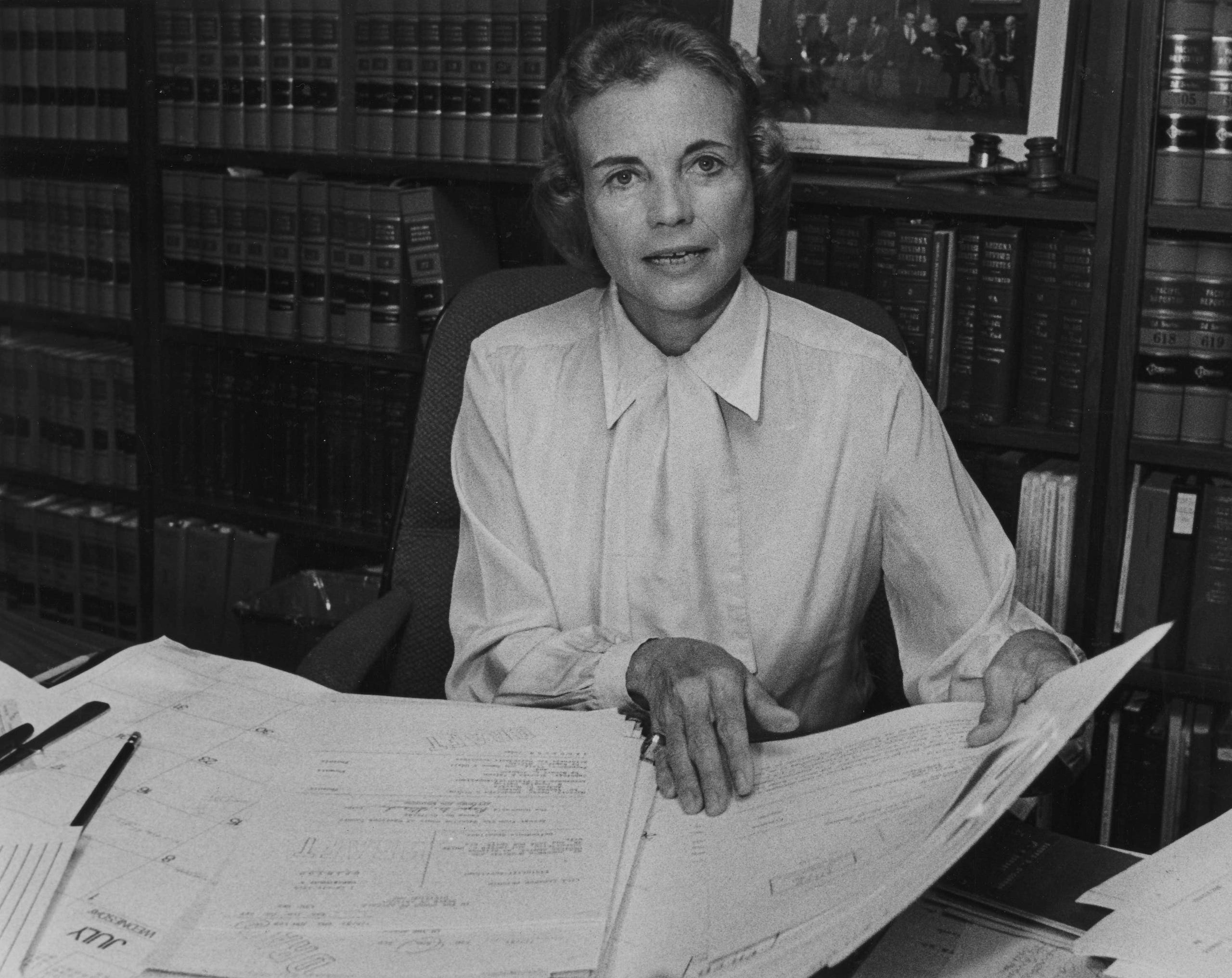 A woman sits at a desk covered with documents and large papers, holding one sheet. She wears a light blouse and is surrounded by shelves of books on civics education, with a framed photo visible behind her.
