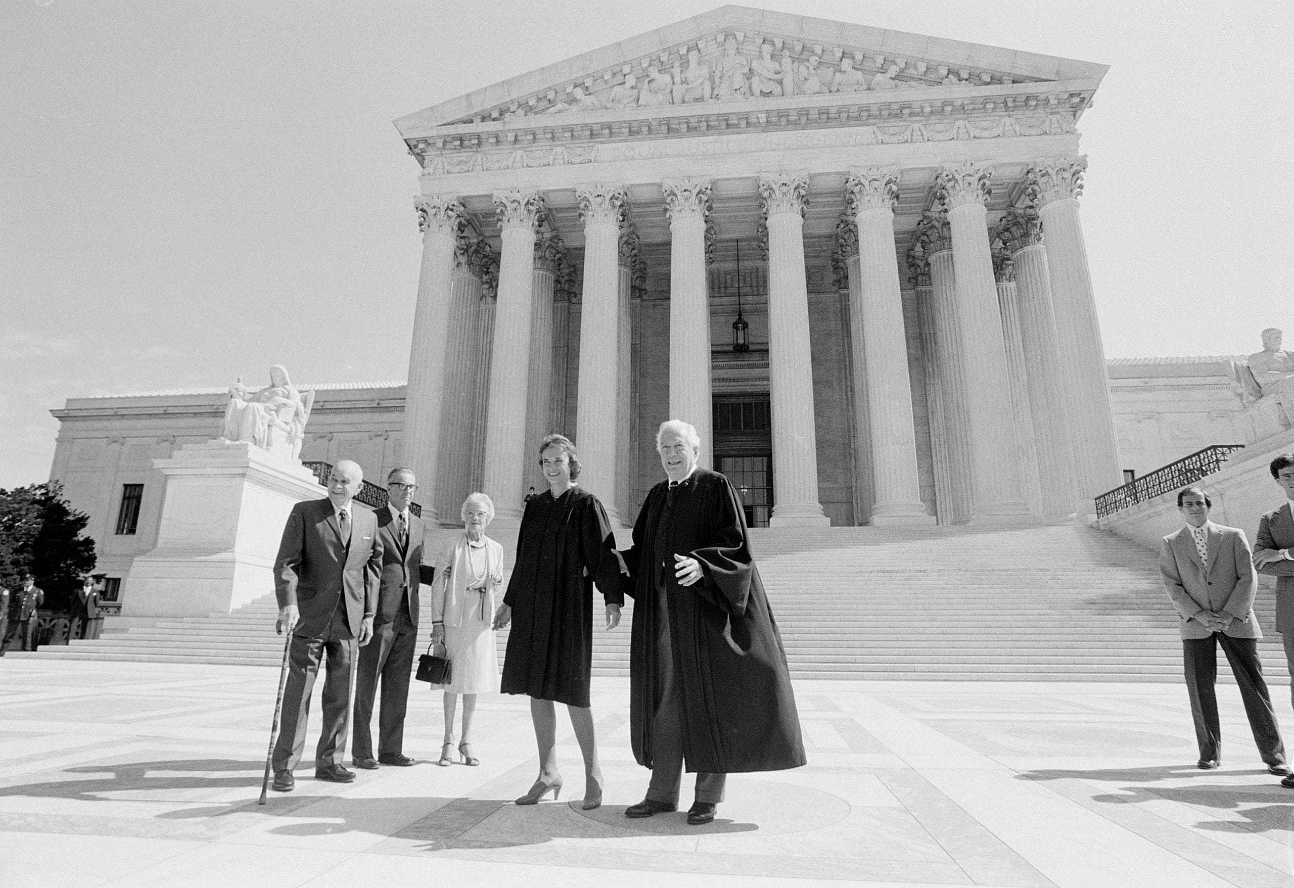 A group of people stand on the steps of a large, neoclassical building with tall columns, likely a courthouse. Two individuals in judicial robes are prominent. Others are dressed in formal attire, and one person uses a cane.
