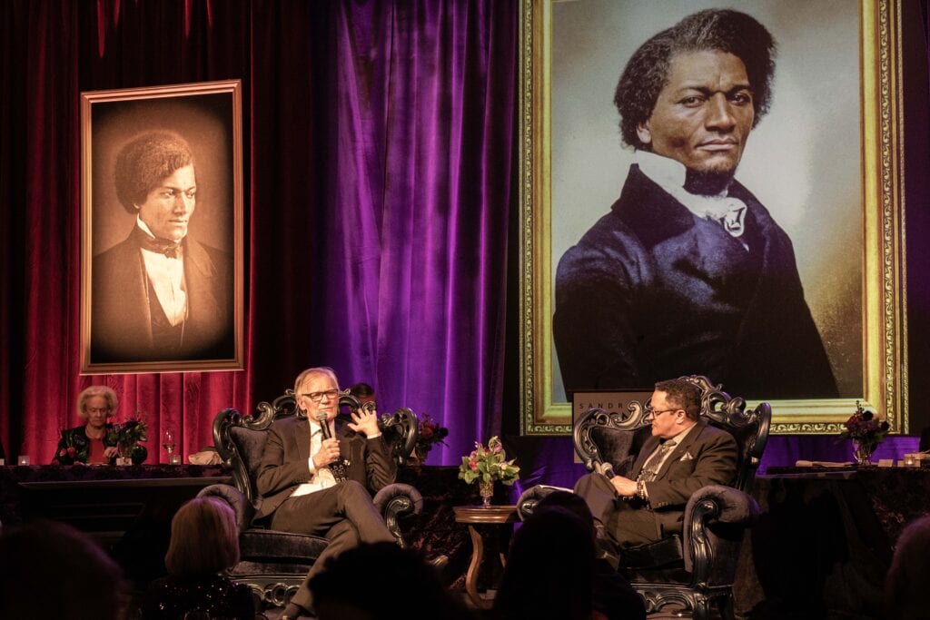 Two people sit onstage in armchairs, engaged in conversation at an event. Behind them are large portraits of Frederick Douglass, set against rich purple curtains. Flowers and a small table are visible on stage. An audience is partially visible in the foreground.