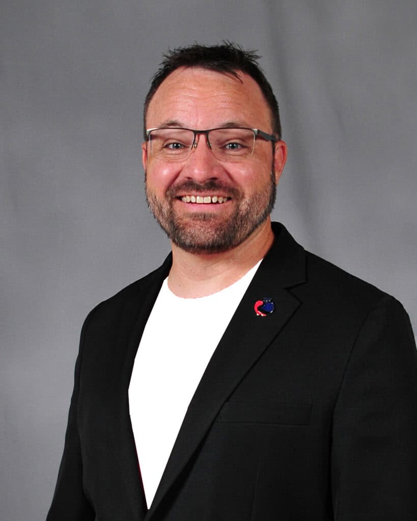 A man with short dark hair and glasses is smiling, wearing a black blazer over a white shirt, standing against a plain gray background.