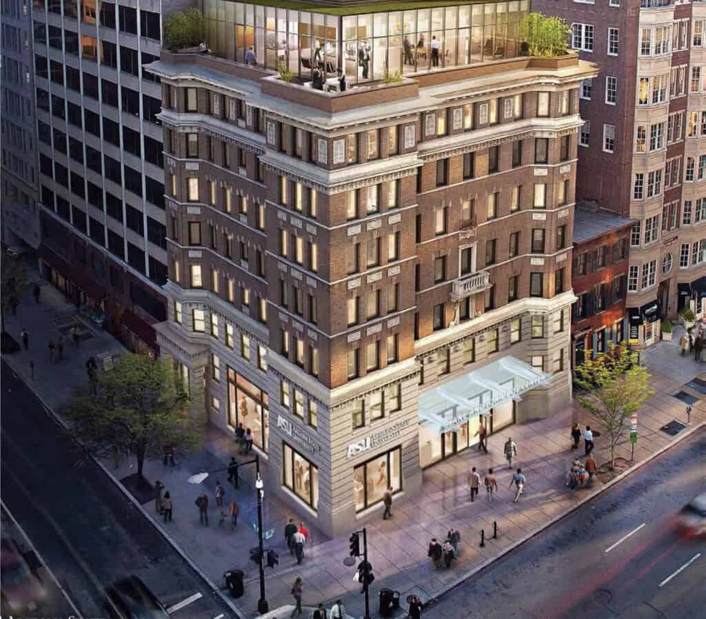 Aerial view of a multi-story brick building with large windows and lit storefronts at street level. People walk by lush trees on the busy urban intersection. The upper floors have balconies and rooftop greenery—contact us for more details.