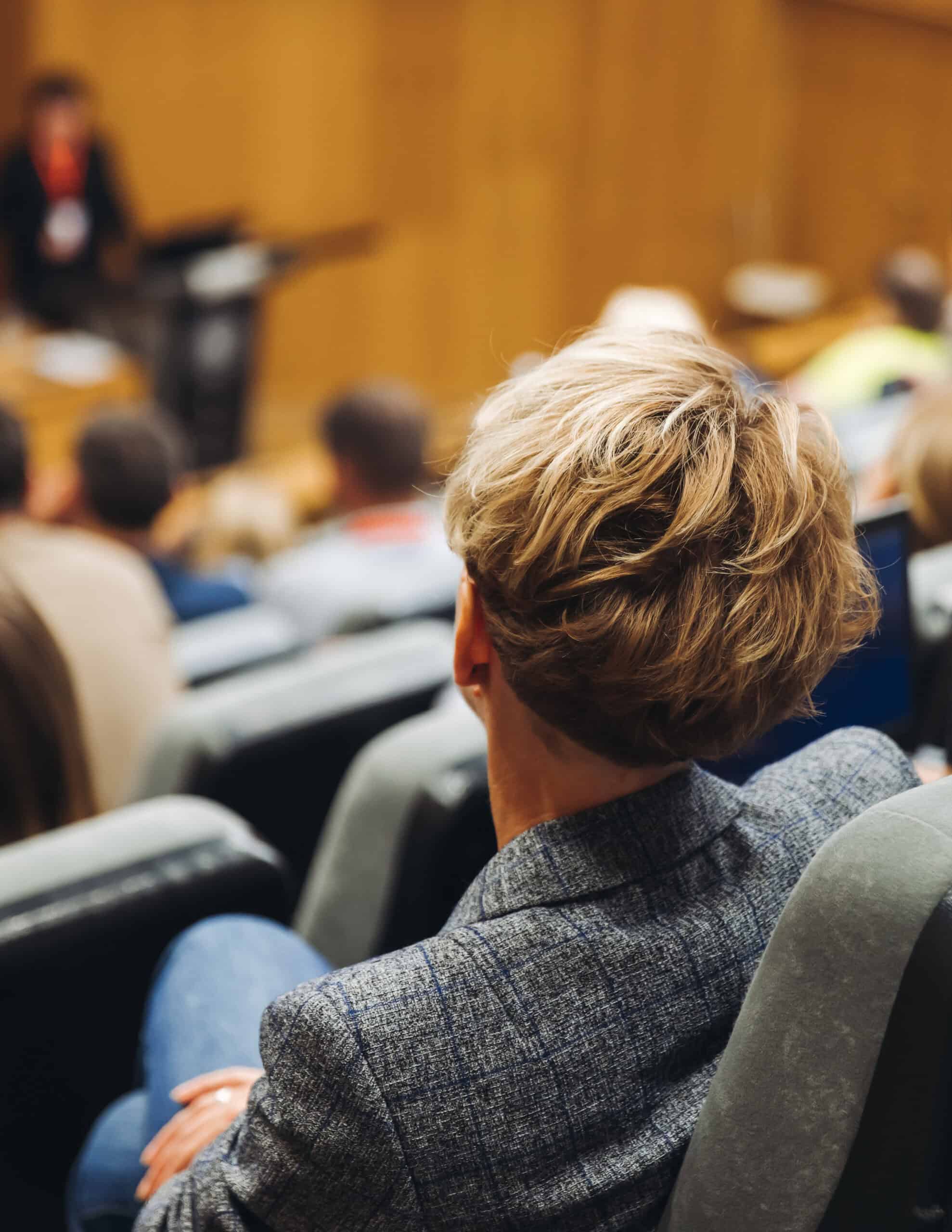 A person with short blonde hair sits in an auditorium, facing a speaker at the front during a Capital Connections event. Rows of attendees are seated, and the setting suggests a lecture or conference.