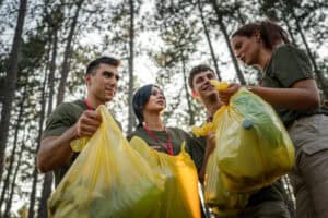group of teenage friends gen Z male and female caucasian men women picking up waste garbage plastic bottles and paper from the forest cleaning up nature in sunny day environmental care ecology concept