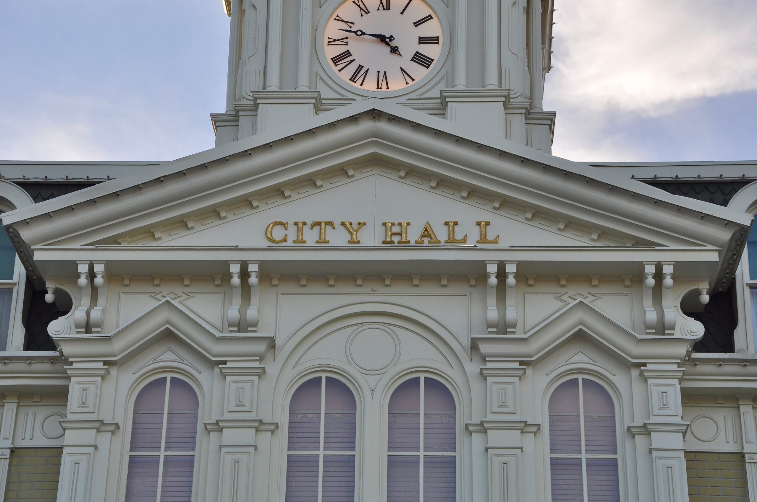 The image shows the upper section of a classic city hall building with a prominent clock and the words "City Hall" engraved above the entrance in gold letters. The architecture features ornate detailing and arched windows.