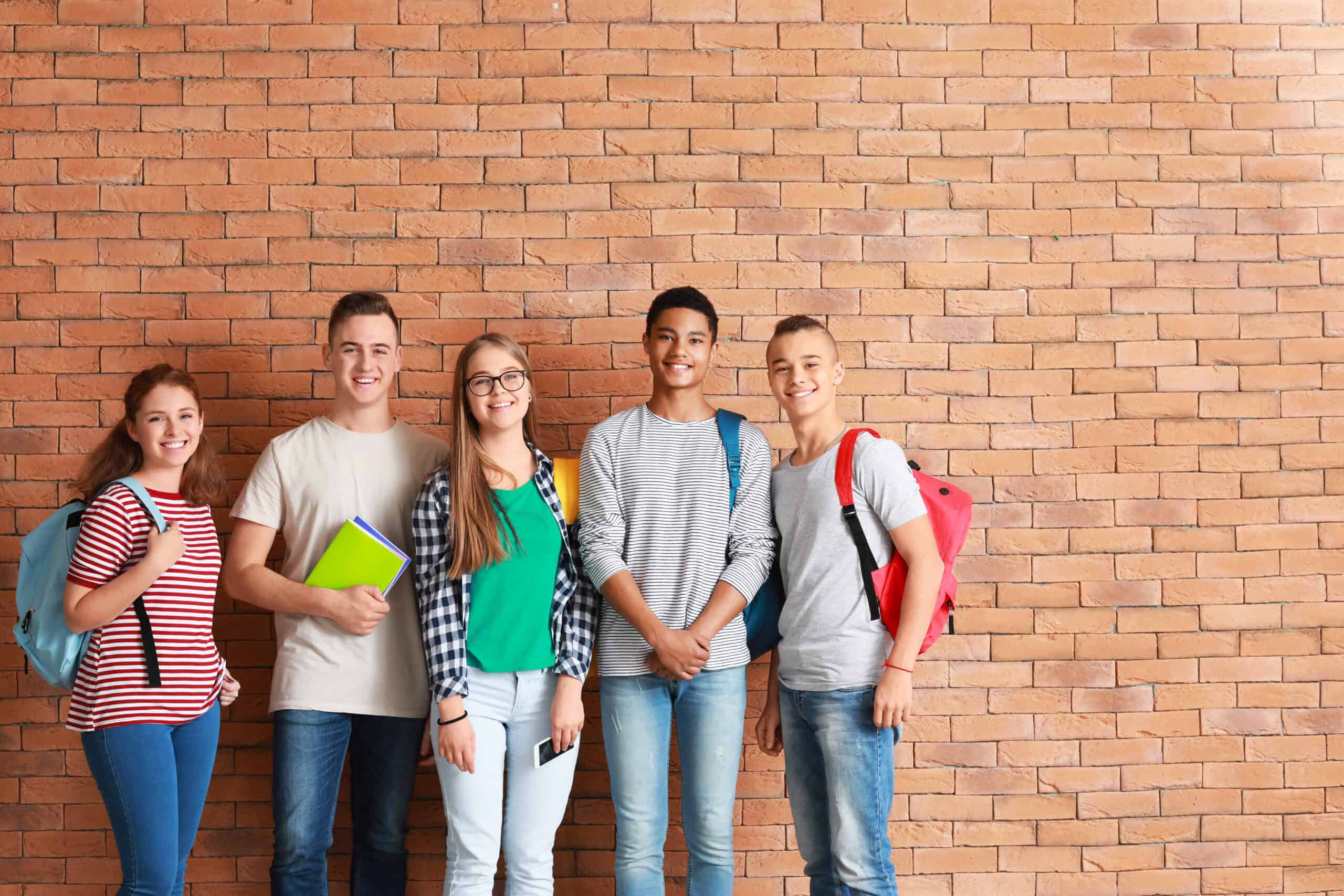 Five smiling teenagers stand in front of a brick wall, each carrying backpacks and books, looking at the camera. Their casual jeans and t-shirts reflect a relaxed atmosphere of student engagement and camaraderie.