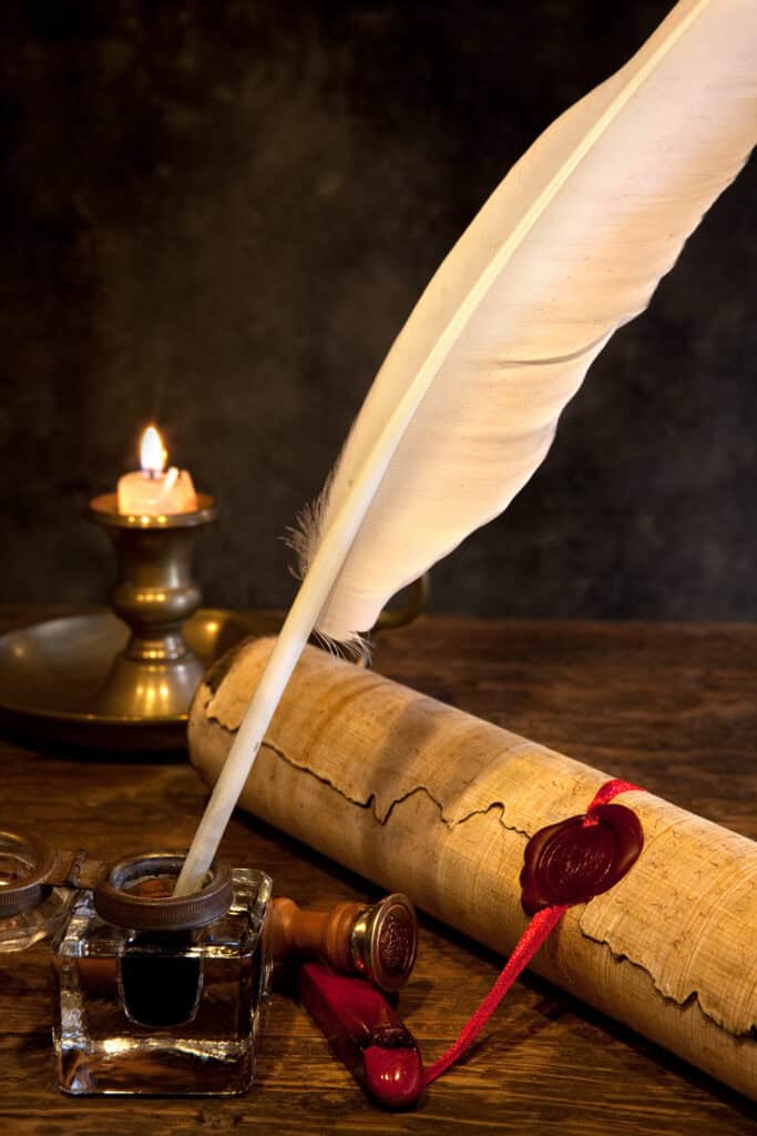 A feather quill in an ink bottle sits next to a rolled parchment sealed with red wax, a wax seal stamp, and a lit candle on a brass holder—an evocative scene that captures the spirit of America's 250th on a wooden surface.