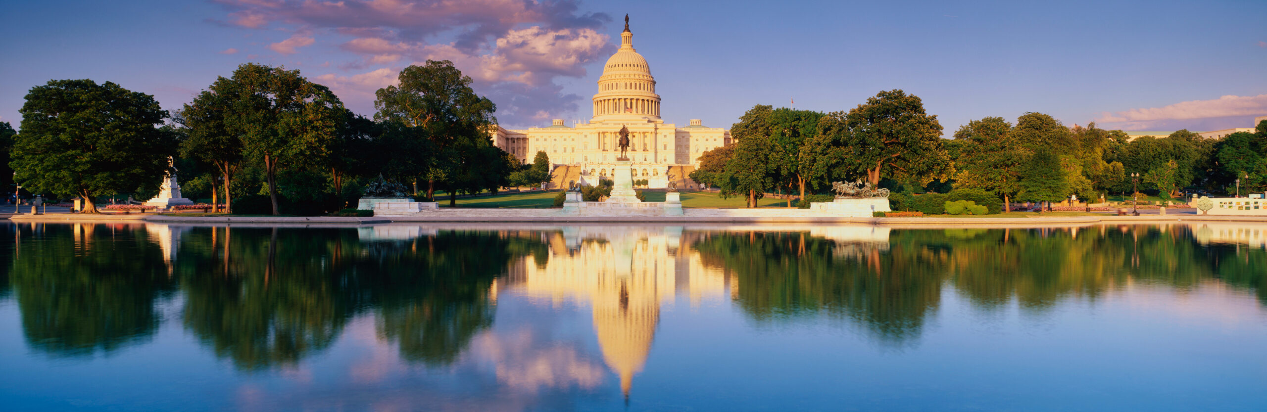 The U.S. Capitol building is seen at sunset with its white dome reflected in a calm, blue pool, surrounded by green trees and a clear sky—an inspiring scene reminiscent of Sandra Day O'Connor’s legacy in American history.