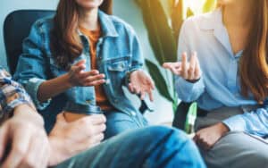 Three people are sitting and having a conversation about must-learn civics. One person holds a coffee cup, while the others gesture as they discuss civics topics in a bright, casually dressed indoor setting.