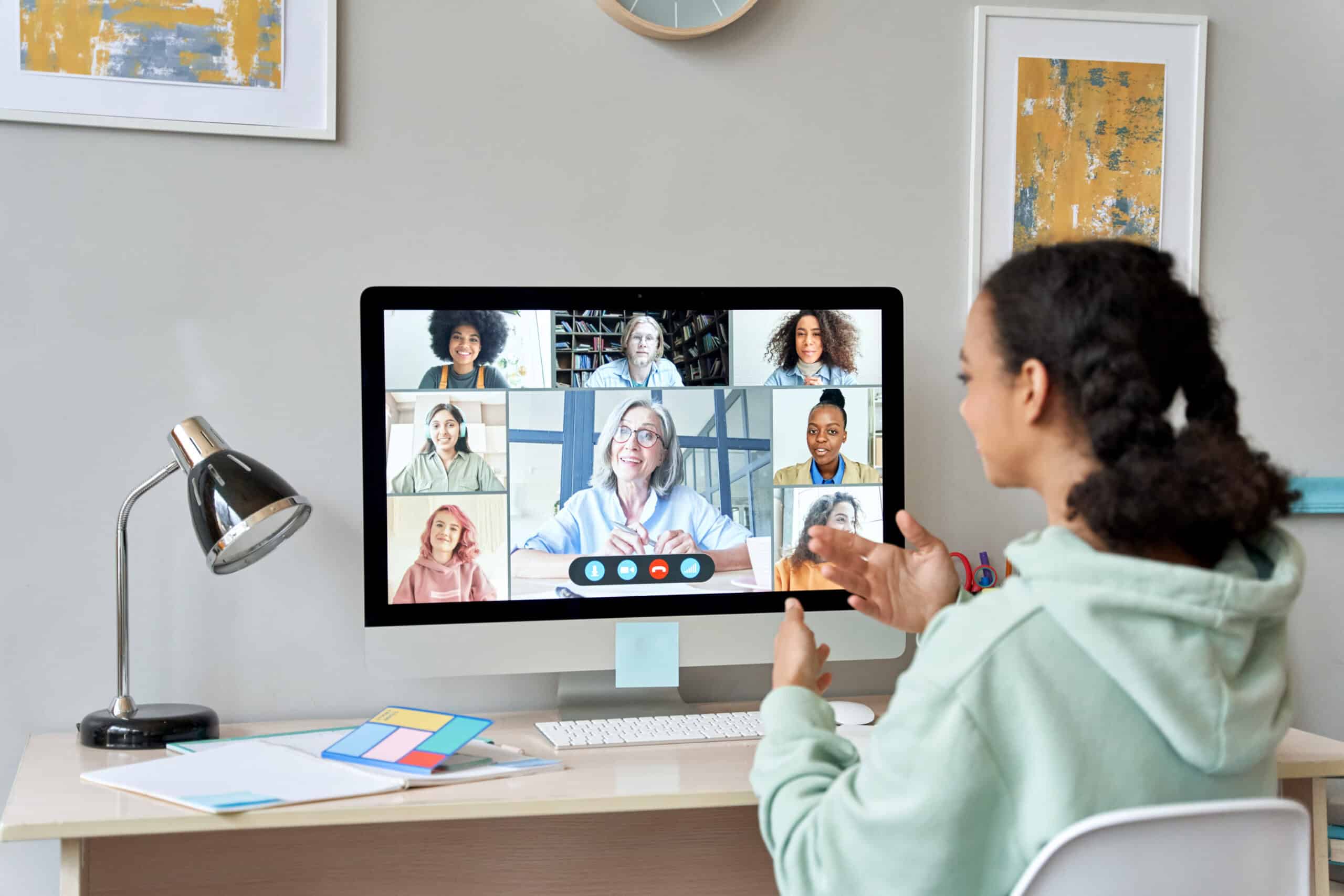 A young woman sits at a desk, joining a video call on her computer with seven diverse ambassadors, all visible in small windows on the monitor. She gestures while speaking, and office supplies are on the desk.