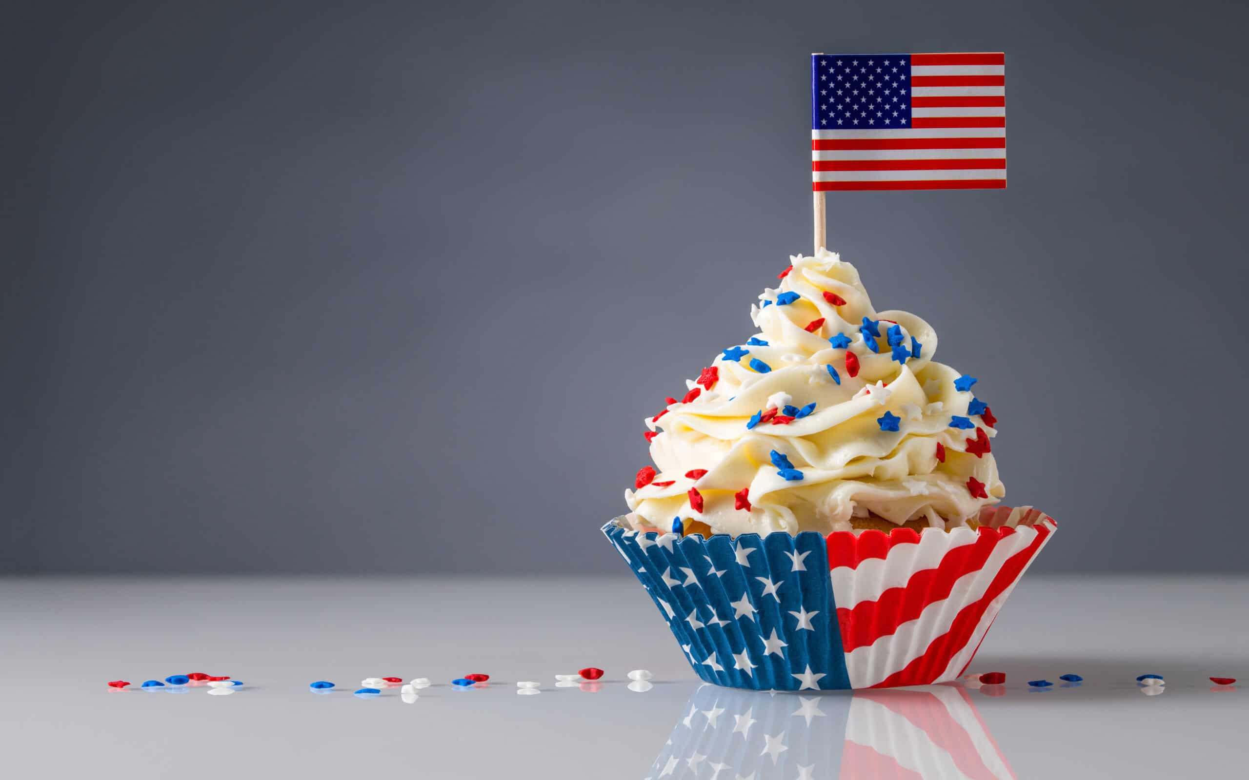 A cupcake with white frosting and red, white, and blue star sprinkles sits in an American flag-patterned wrapper. Topped with a small U.S. flag, it's a festive treat perfect for celebrating America's 250th anniversary.