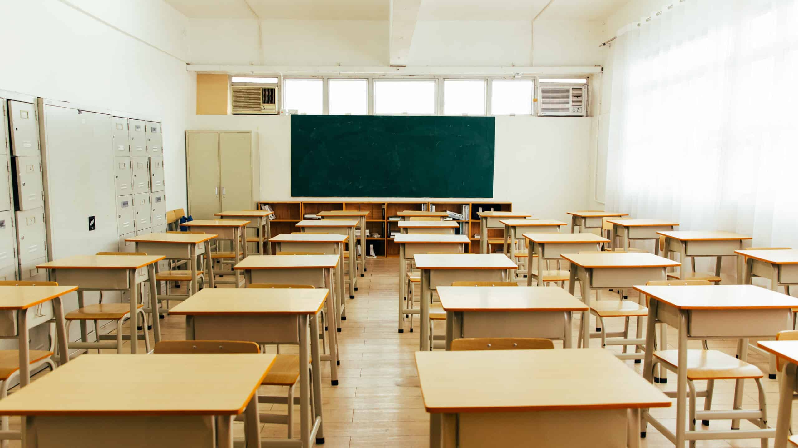 Empty classroom with multiple wooden desks and chairs arranged in rows, a large green chalkboard at the front—ideal for teaching civics—lockers on the left wall, and sunlight streaming through windows on the right.