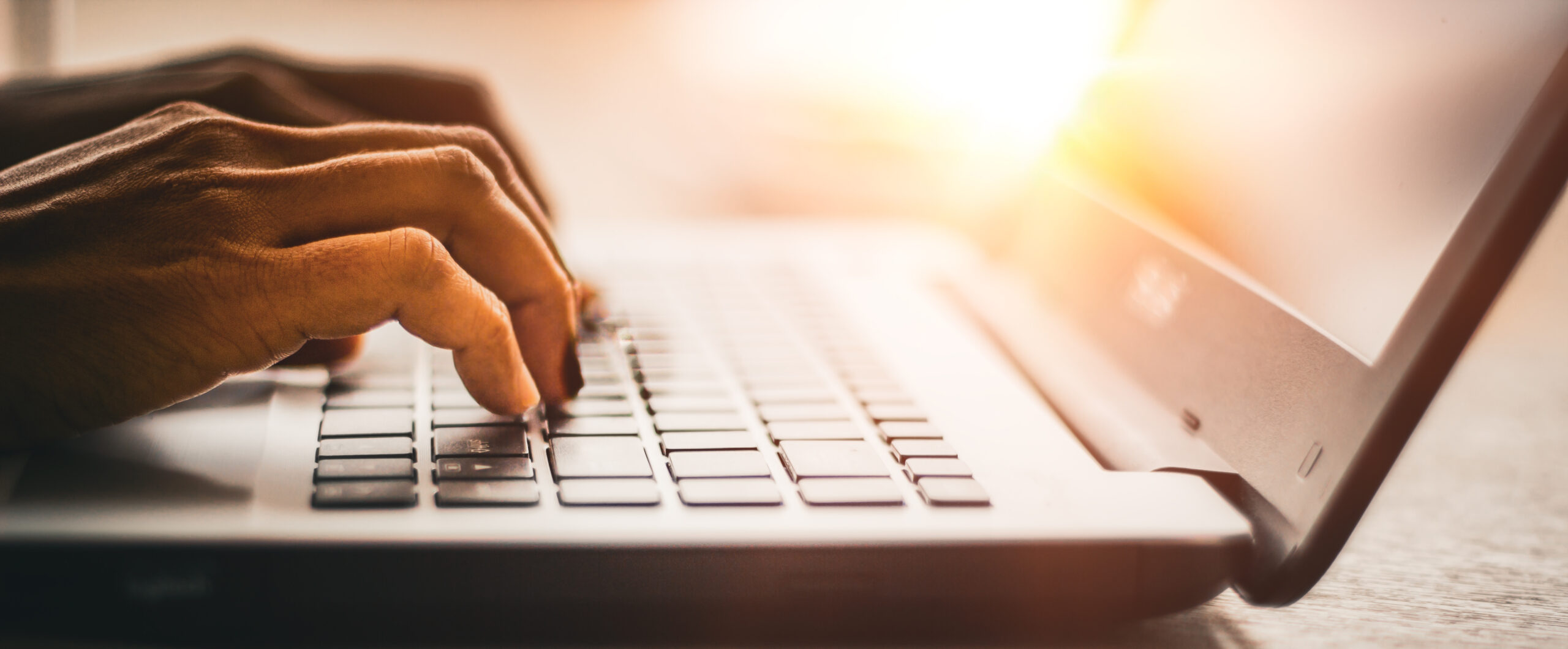 Close-up of hands typing on a laptop keyboard with bright sunlight shining in the background, creating a warm and focused atmosphere.