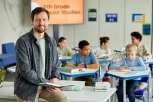A teacher holding a book smiles at the camera while sitting on a desk in a classroom with a "6th Grade Social Studies" sign, where students work in the background—highlighting the importance of Must Learn Civics for young minds.