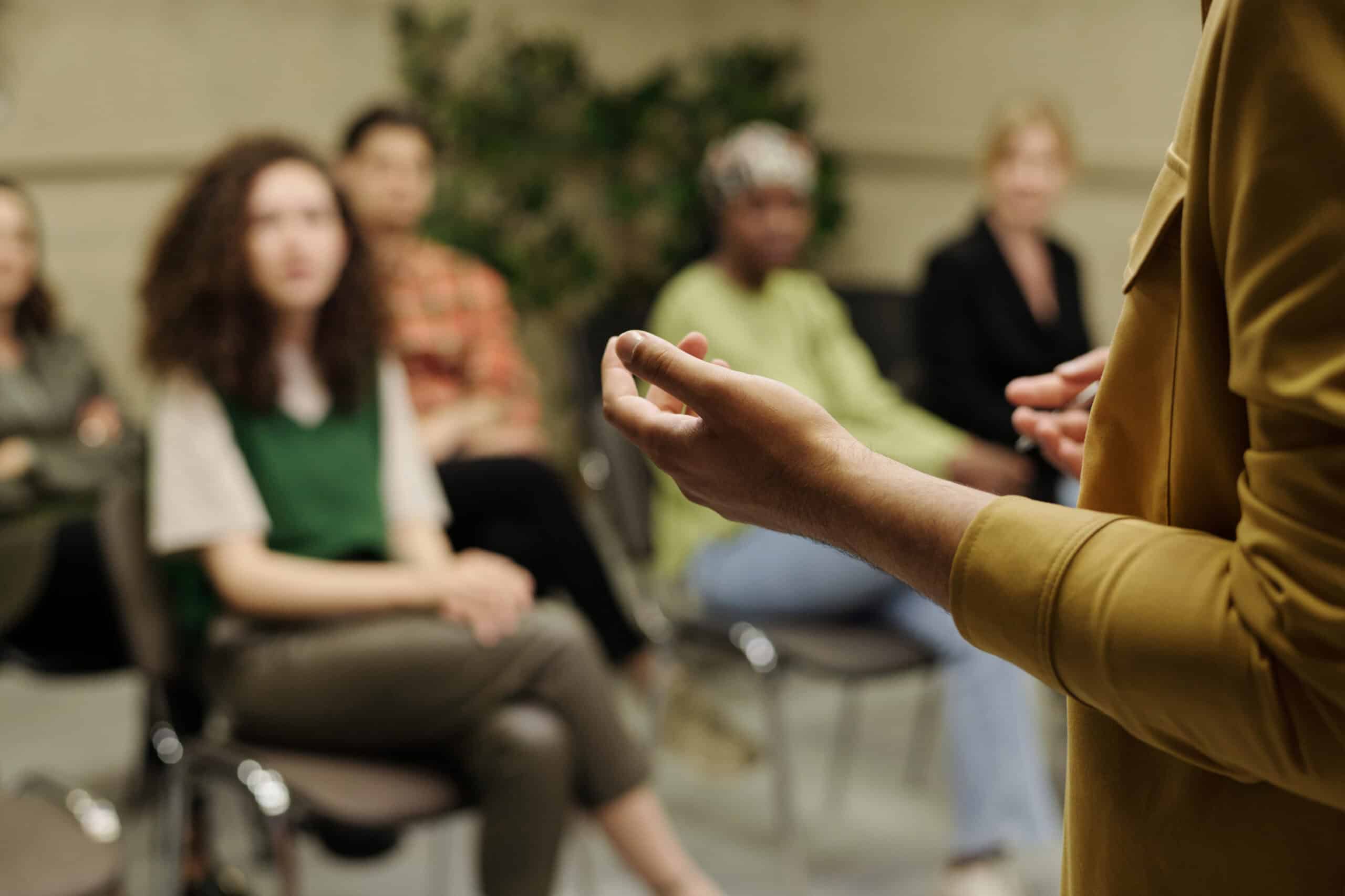 A person in a yellow shirt speaks to a seated group of four, fostering student engagement as they listen attentively in a casual, indoor setting. The focus is on the speaker’s hand in the foreground.