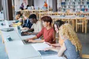 A group of diverse students sits at a long table in a library, studying together and showing strong student engagement. Some use laptops while others write in notebooks, surrounded by shelves of books and more desks in the background.