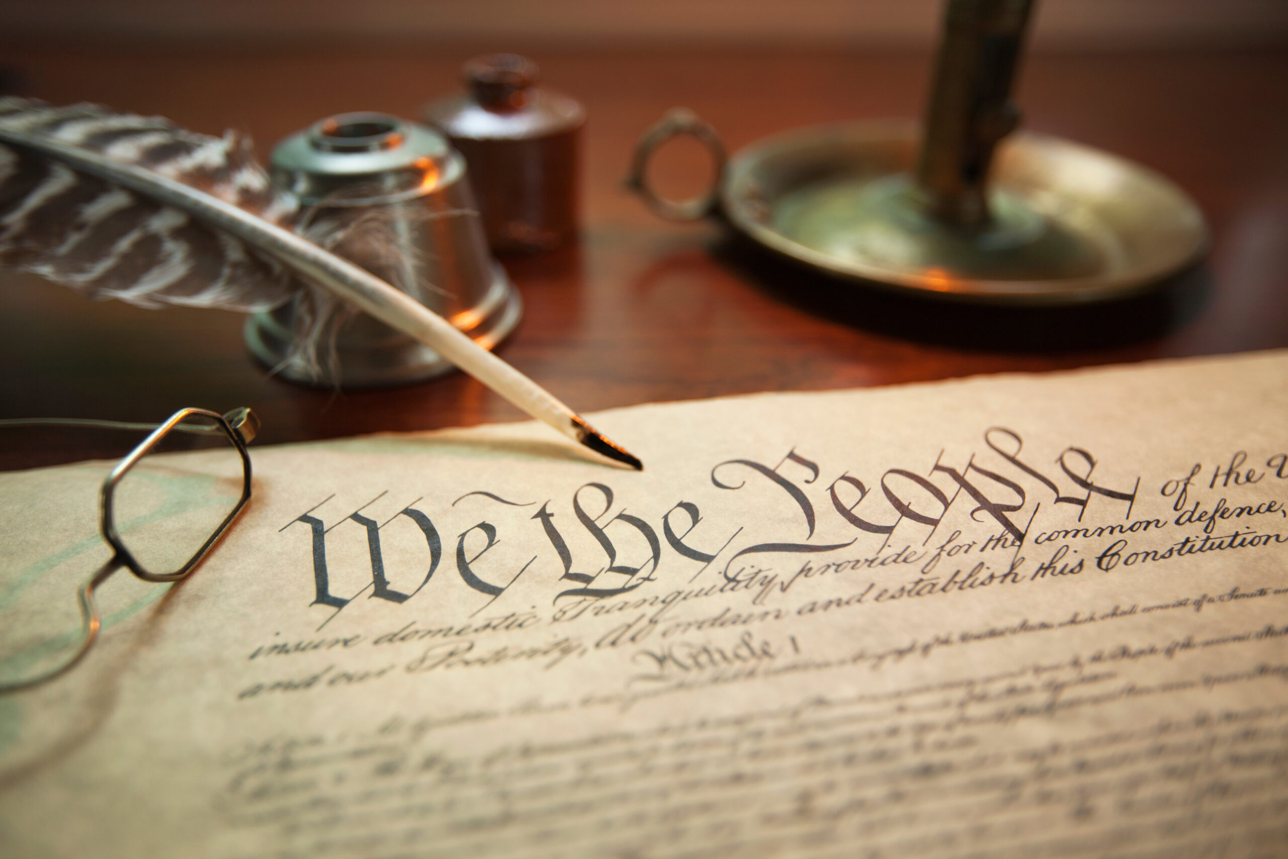 A close-up of the U.S. Constitution’s preamble, featuring “We the People,” with a quill pen, ink bottle, eyeglasses, and a brass candle holder on a wooden desk—evoking the spirit of America’s 250th anniversary.