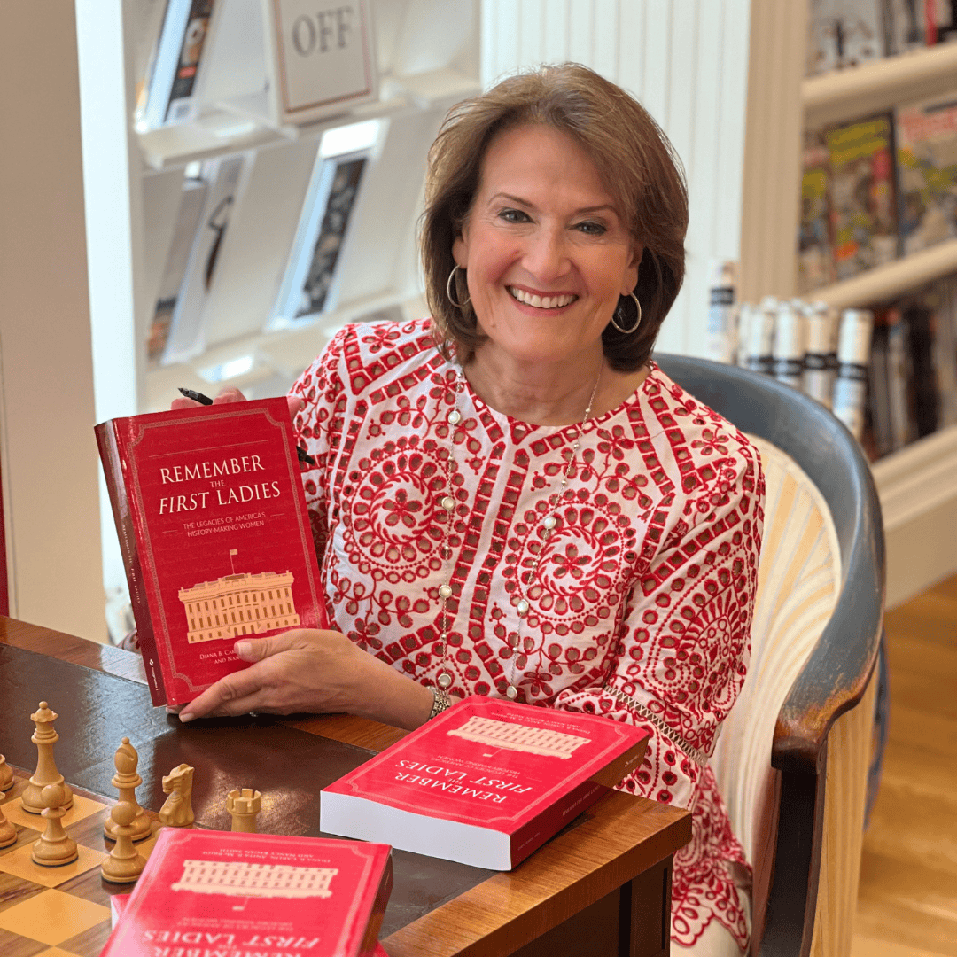 A woman smiling and sitting at a table with chess pieces, holding a red book titled "Remember the First Ladies," celebrates women in leadership. More copies of the book are on the table, with bookshelves and magazines in the background.