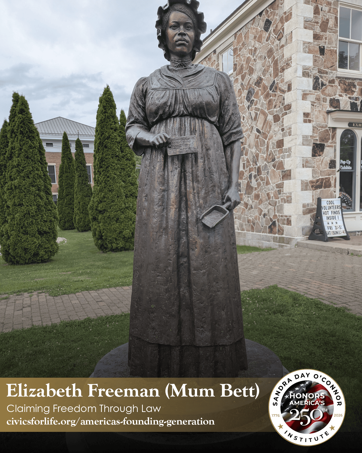 A bronze statue of Elizabeth Freeman (Mum Bett), known as The Citizen, stands outdoors on a brick walkway, with tall green shrubs and a stone building with large windows in the background.