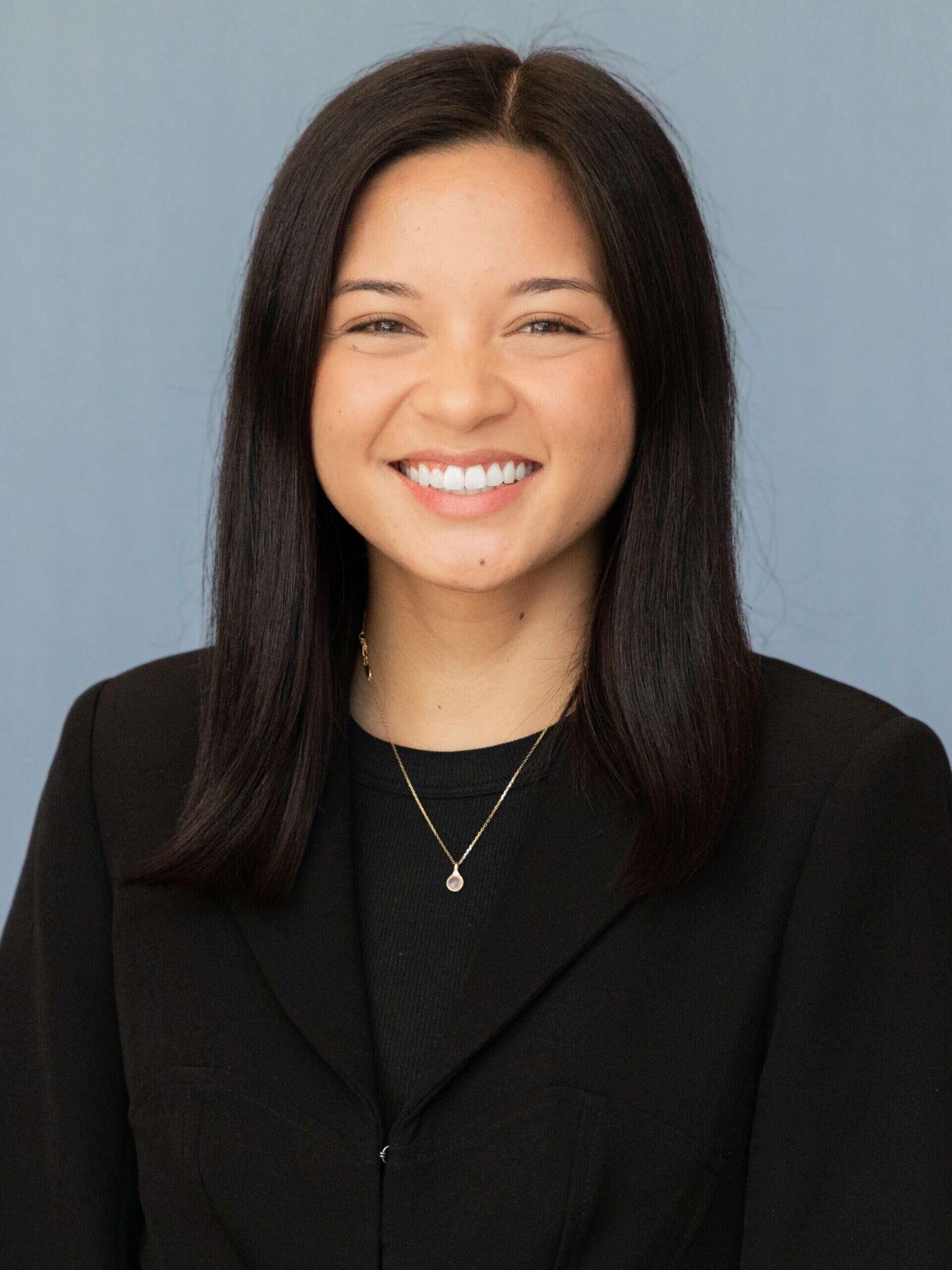 A woman with straight, dark hair wearing a black blazer and a gold necklace smiles at the camera in front of a plain light blue background, embodying the professional spirit of Capital Connections.