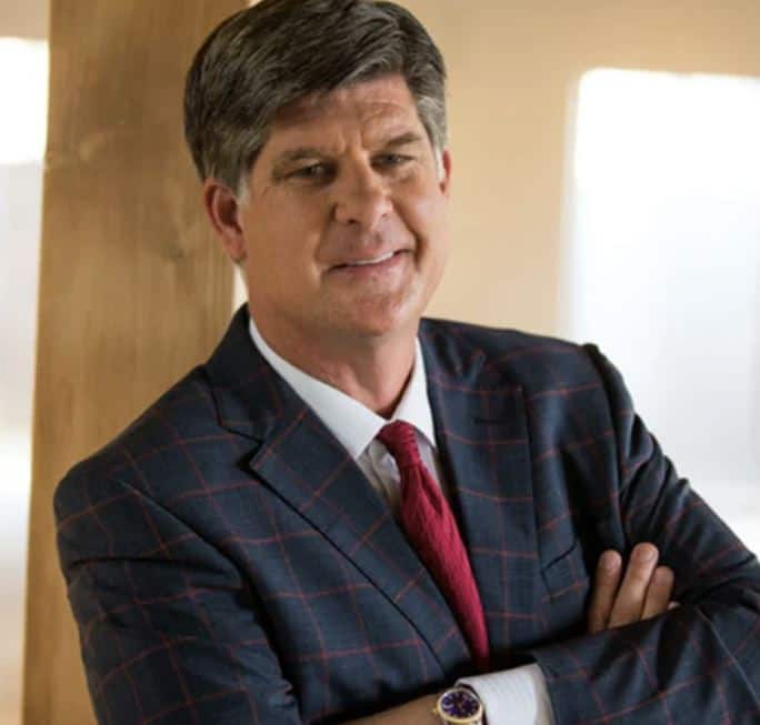 A middle-aged man with short dark hair, wearing a checkered navy suit, white shirt, and red tie, stands with arms crossed and smiles in a softly lit indoor setting.