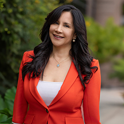 A woman with long dark hair, wearing a bright orange blazer over a white top, smiles while standing outdoors with greenery in the background.