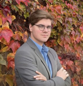 A person with glasses and light brown hair, wearing a gray blazer and blue shirt, stands with arms crossed in front of a wall covered in colorful autumn ivy leaves.