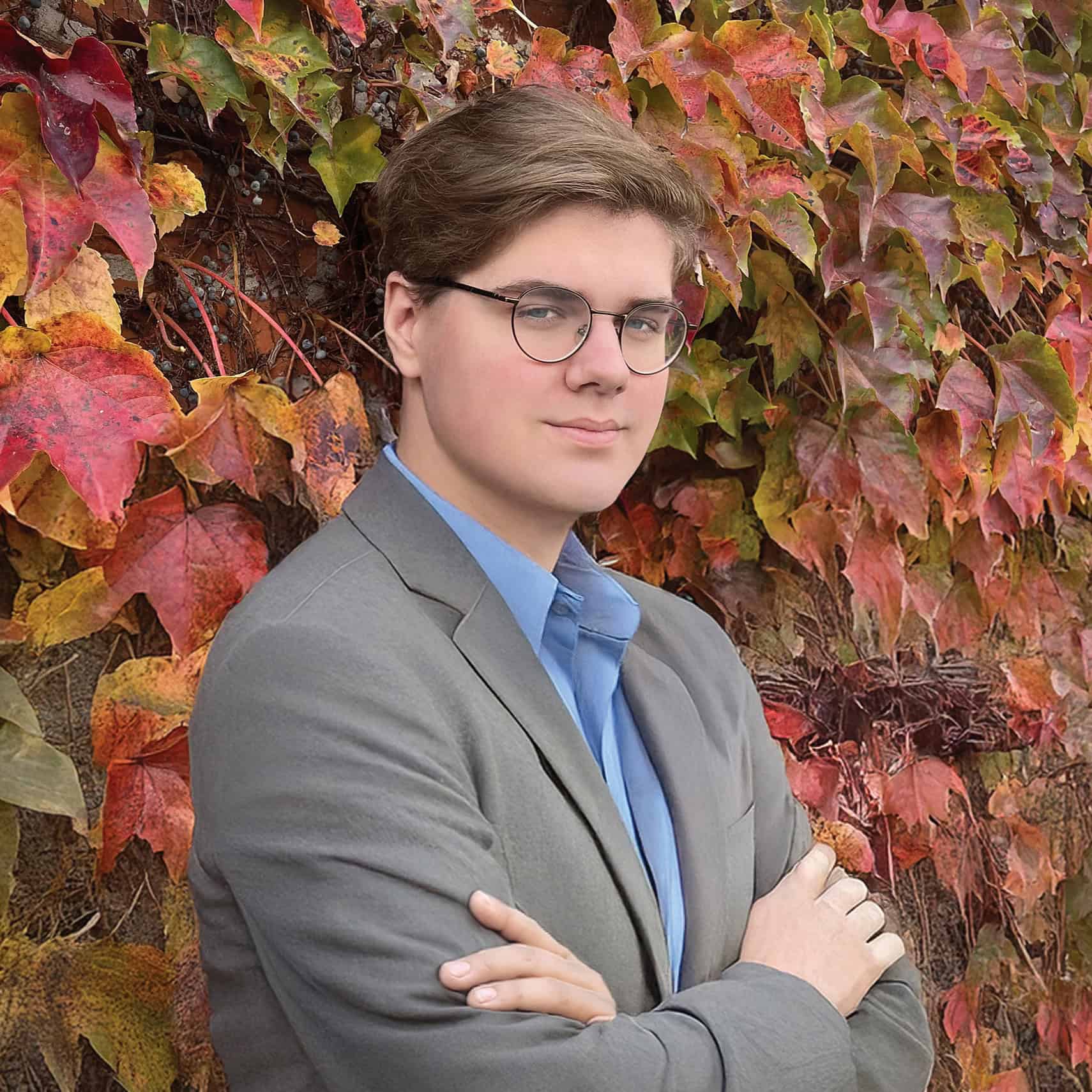 A young man with glasses and short brown hair, wearing a gray blazer and blue shirt, stands with arms crossed in front of a wall covered in autumn-colored ivy leaves, embodying the confidence often seen in scholarship winners.
