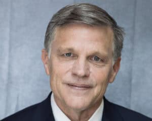 A middle-aged man with short, gray hair and blue eyes, wearing a dark suit and white shirt, smiles slightly in front of a plain gray background, as if ready to share a history dinner story.