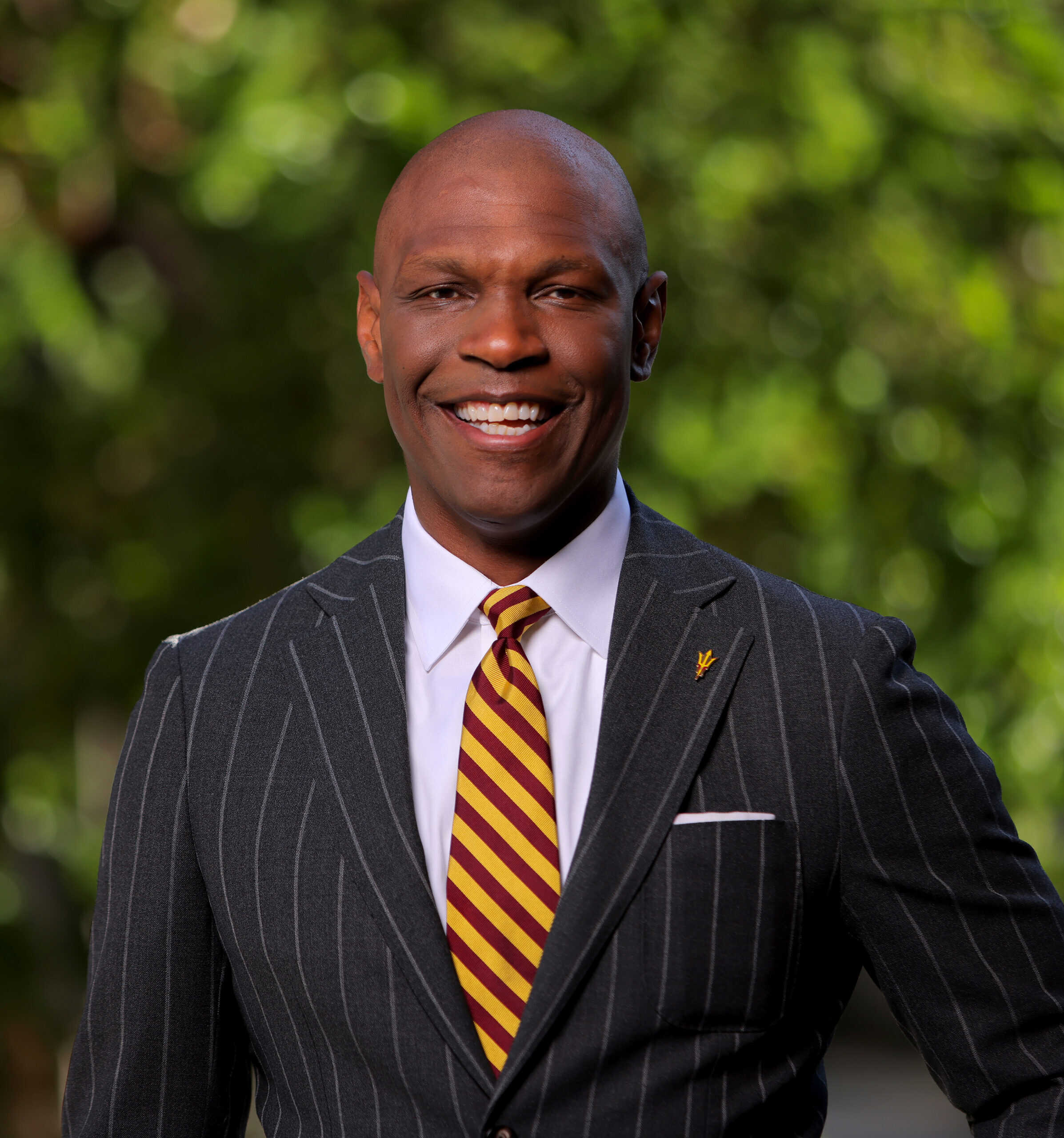A smiling man in a dark pinstripe suit and a maroon and gold striped tie stands outdoors in front of a leafy green background, representing his 501(c)(3) nonprofit organization with pride.