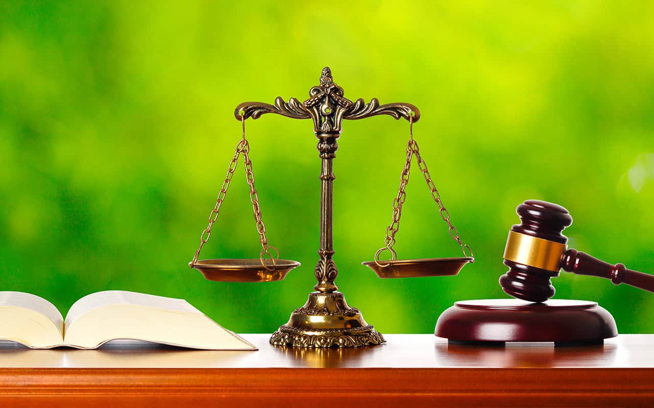 A brass balance scale, an open book, and a wooden judge's gavel rest on a wooden surface against a green, blurred background.